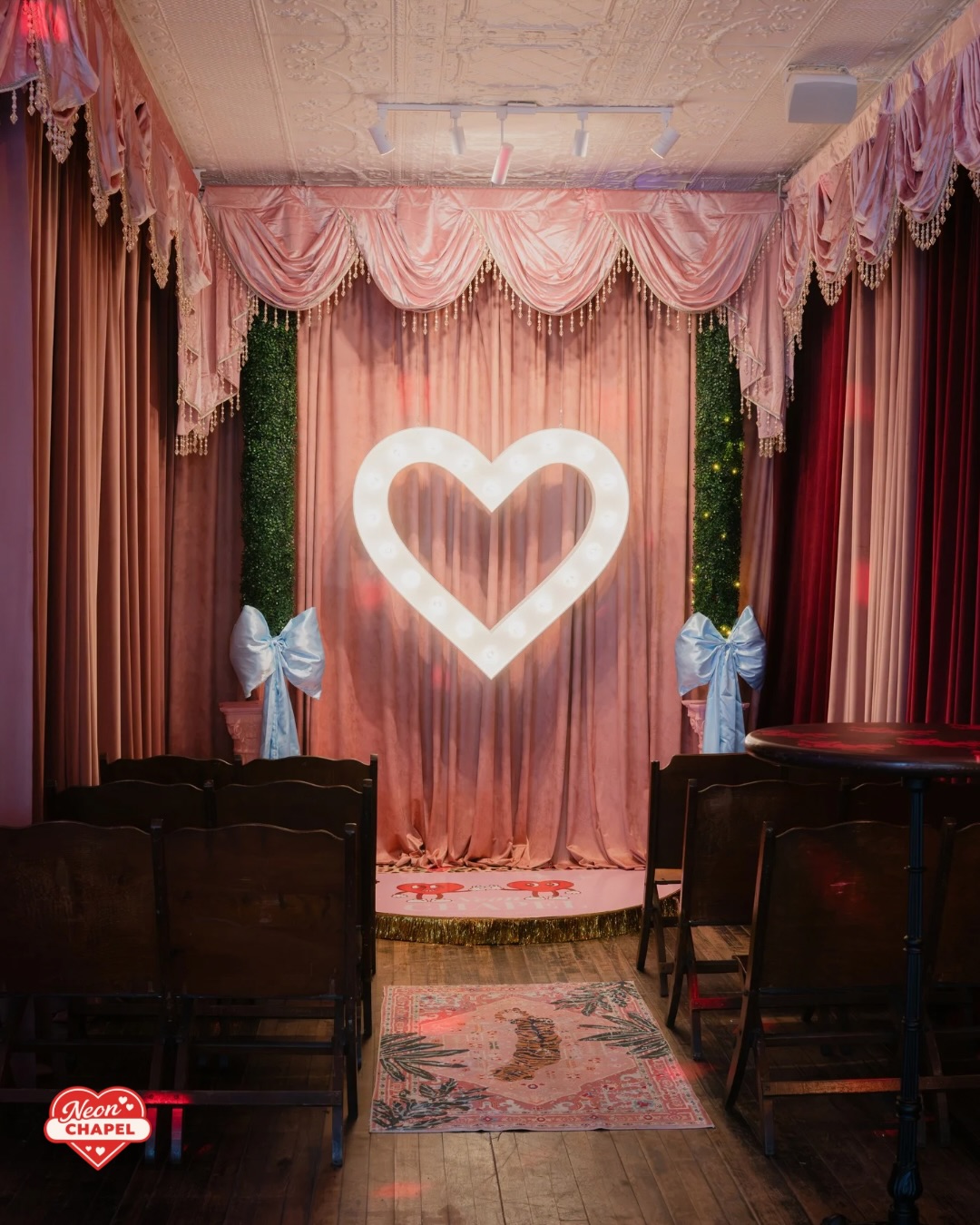 The Neon Chapel ceremony room: a glowing marquee heart against blush velvet curtains, with wooden pews and a tiger rug down the aisle