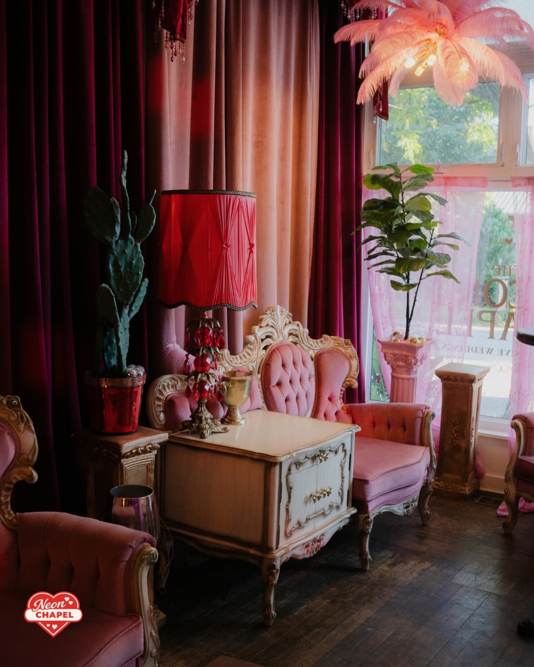 Cozy corner with a pink feather chandelier, gilded tufted loveseat and a red beaded lamp by the window