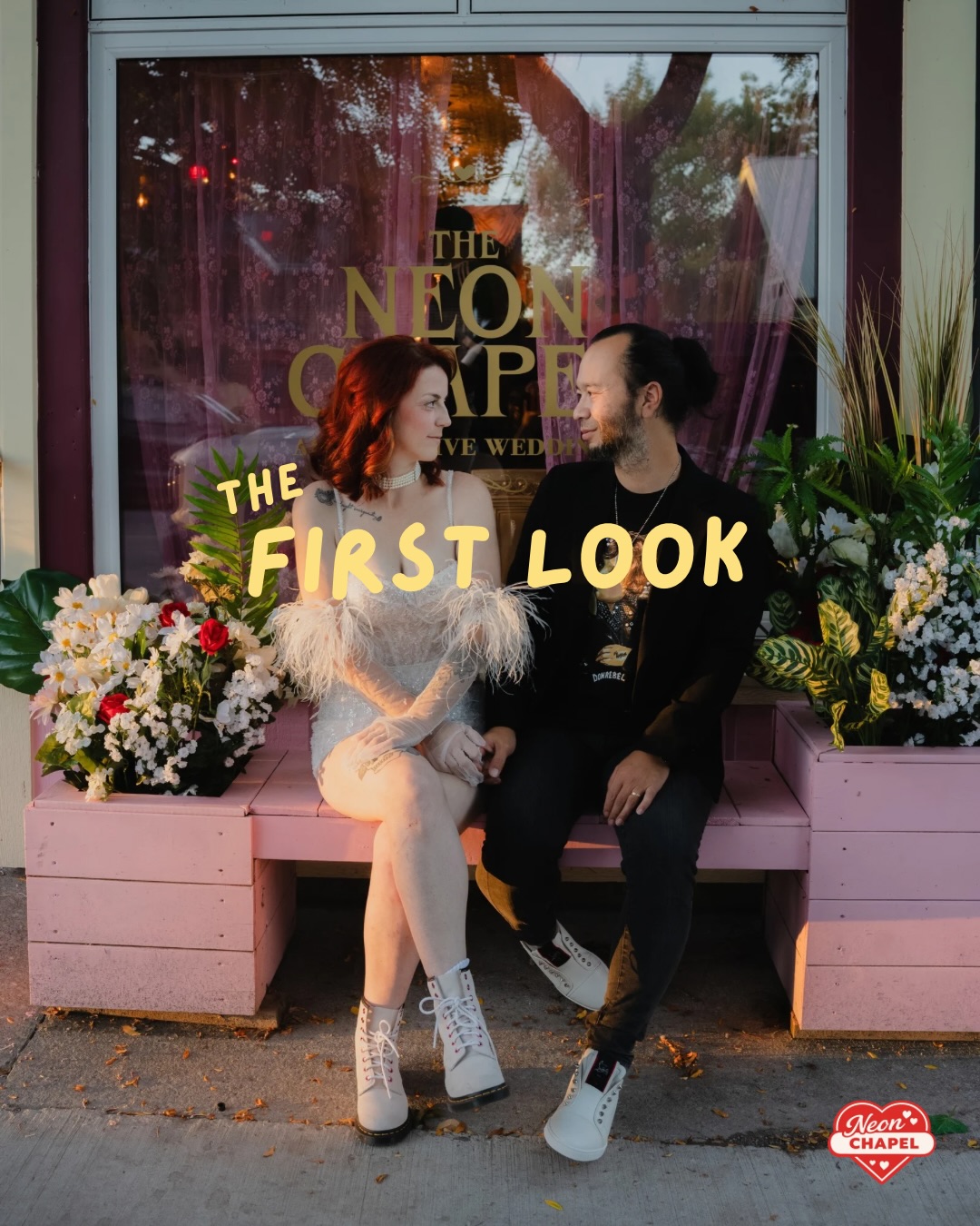 A newlywed couple sitting on a pink bench outside The Neon Chapel window on Wellington Main Street